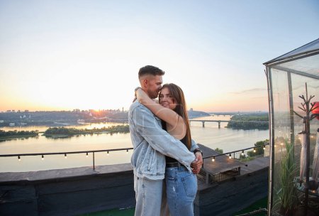 A young couple kisses and embraces during a romantic rooftop date, with a cityscape backdrop at sunset.
