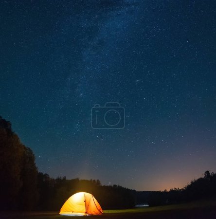Foto de Una impresionante foto nocturna con una carpa amarilla brillando desde el interior, rodeada por un bosque oscuro, y la Vía Láctea y las estrellas brillando en el cielo. - Imagen libre de derechos