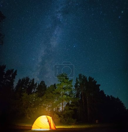An awe-inspiring night shot capturing a yellow tent lit up from within, and the awe-inspiring Milky Way and stars in the dark night sky.