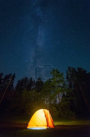 A stunning nighttime photo featuring a yellow tent glowing from within, surrounded by a dark forest, and the Milky Way and stars glittering in the sky.