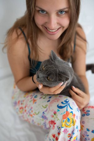 Téléchargez les photos : Jeune femme souriante avec son chat à poil court britannique dans son lit - en image libre de droit