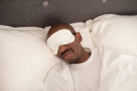 Photo for Young black guy  sleeping with a blindfold mask covering his eyes lying in a bed - Royalty Free Image
