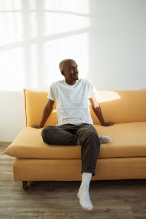 Photo for Young black man relaxing at home on the yellow couch in a room lit with sunshine - Royalty Free Image