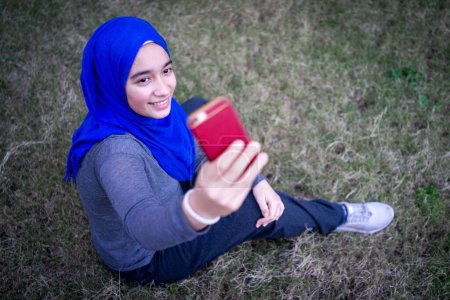 Téléchargez les photos : Musulmane jeune femme assise sur l'herbe ang prendre selfie seul - en image libre de droit