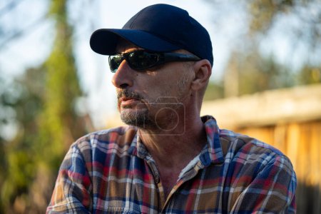 Téléchargez les photos : Homme mature auto assuré en chemise à carreaux croisant les bras regardant la caméra debout dans le village. - en image libre de droit