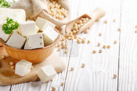 Tofu au soja caillé dans un bol en bois et dans un sac de chanvre sur une table de cuisine en bois blanc. Substitut non laitier au fromage. Place pour le texte
