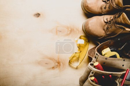 Construction concept background of toolbelt yellow boots goggles ant tools on plywood flat lay