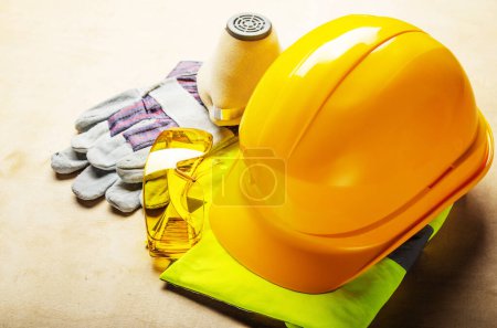 Gelbe Plastikmütze, Brille, Atemschutzmaske und Schutzhandschuhe liegen auf Sperrholz. Sicheres Arbeitsschutzkonzept
