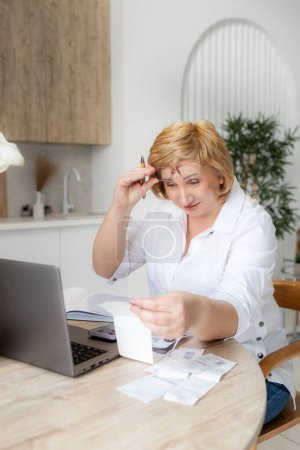 Senior mature business woman holding paper bill using laptop. Old lady managing account finance, calculating money budget tax, planning banking loan debt pension payment sit at home kitchen table.