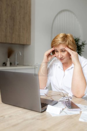 Senior mature business woman holding paper bill using laptop. Old lady managing account finance, calculating money budget tax, planning banking loan debt pension payment sit at home kitchen table.