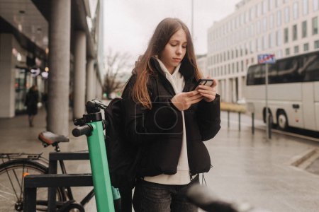 School age girl renting an electric scooter or bicycle using smartphone, making contactless payment through mobile app. Bicycle rental in the city in the autumn-winter season.