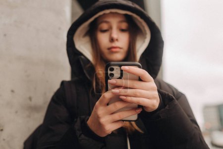 Half length portrait of girl in casual wear holding smartphone for blogging in the city in the autumn-winter season. Teen surfing the internet outdoors.