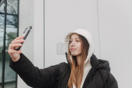 Photo for Half length portrait of girl in casual wear holding smartphone for blogging in the city in the autumn-winter season. Teen surfing the internet outdoors. - Royalty Free Image