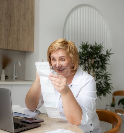 Photo for Senior mature business woman holding paper bill using laptop. Old lady managing account finance, calculating money budget tax, planning banking loan debt pension payment sit at home kitchen table. - Royalty Free Image