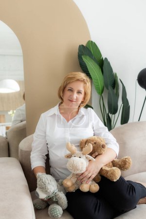 Happy grandmother and teddy bear toy. Cheerful and smiling grandmother sitting on sofa at home.