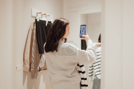 Foto de Chica adolescente coreana se toma una selfie en ropa nueva en el espejo y se la envía a su amiga por teléfono en un centro comercial. Retail y consumismo. Promoción de venta y concepto de compras. Parte de una serie. - Imagen libre de derechos