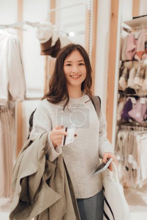 Foto de Chica adolescente coreana tiene una tarjeta de crédito en sus manos y quiere pagar por ropa nueva en un centro comercial. Retail y consumismo. Promoción de venta y concepto de compras. Parte de una serie. - Imagen libre de derechos