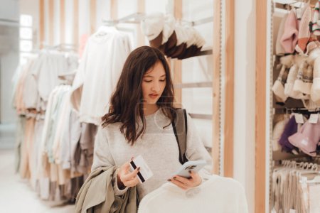 Photo for Korean teenage girl holds a credit card in her hands and wants to pay for new clothes in a shopping mall. Retail and consumerism. Sale promotion and shopping concept. Part of a series. - Royalty Free Image