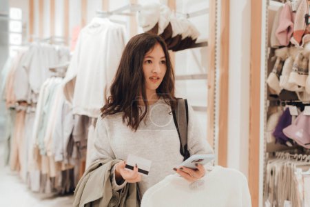 Foto de Chica adolescente coreana tiene una tarjeta de crédito en sus manos y quiere pagar por ropa nueva en un centro comercial. Retail y consumismo. Promoción de venta y concepto de compras. Parte de una serie. - Imagen libre de derechos