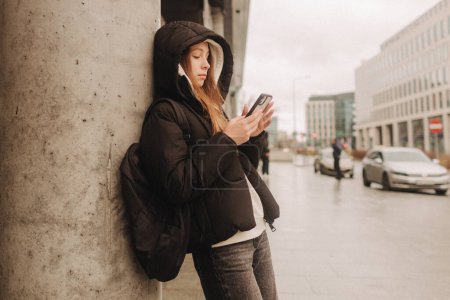 Foto de Retrato de media longitud de una chica en ropa casual sosteniendo un smartphone para bloguear en la ciudad en la temporada otoño-invierno. Adolescente navegando por Internet al aire libre. - Imagen libre de derechos