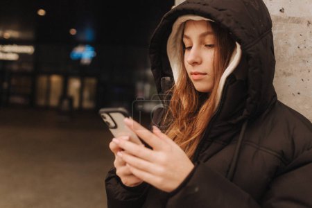 Half length portrait of girl in casual wear holding smartphone for blogging in the city in the autumn-winter season. Teen surfing the internet outdoors.