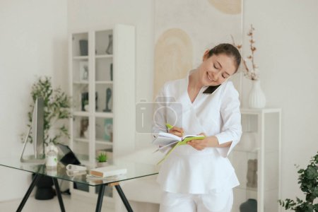 Medicine, healthcare and people concept - Family doctor consults a patient by phone and makes notes, searching for symptoms, online consultation. Woman therapist in white uniform in the clinic office.