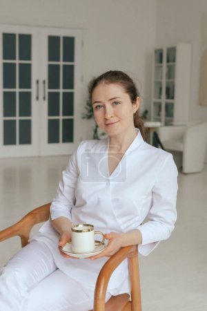 Medicine, healthcare and people concept - Friendly female gp physician doctor in white uniform drinking coffee tea having breakfast while working in the clinic office. Part of a series.