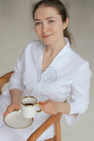 Medicine, healthcare and people concept - Friendly female gp physician doctor in white uniform drinking coffee tea having breakfast while working in the clinic office. Part of a series.