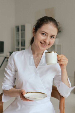 Médecine, soins de santé et concept de personnes médecin gp femme amicale médecin en uniforme blanc boire du thé au café prendre le petit déjeuner tout en travaillant dans le bureau de la clinique. Partie d'une série.