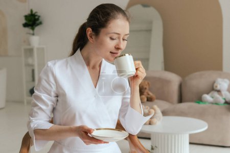 Photo for Medicine, healthcare and people concept - Friendly female gp physician doctor in white uniform drinking coffee tea having breakfast while working in the clinic office. Part of a series. - Royalty Free Image