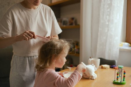Happy single mother doing her daughter's hair while having breakfast. Woman combs the hair of a little girl. The concept of morning routine, single parenthood and preparation for school.