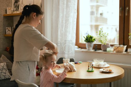 Happy single mother doing her daughter's hair while having breakfast. Woman combs the hair of a little girl. The concept of morning routine, single parenthood and preparation for school.