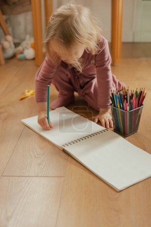 Portrait of a happy child drawing with colored pencils in an album on the floor. Adorable two year old girl in the playroom. Alpha generation concept.
