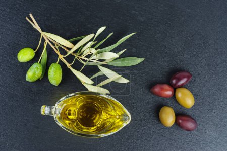 Foto de Delicioso aceite de oliva, aceitunas negras y verdes con hojas, sobre fondo de pizarra de piedra negra - Imagen libre de derechos