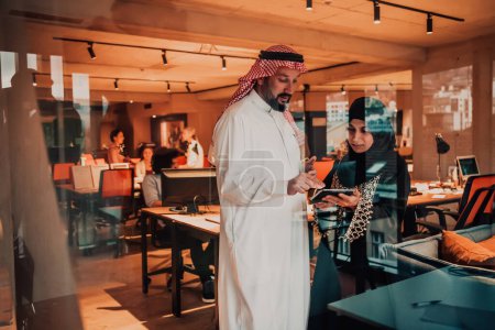 Foto de Foto de retrato del líder árabe hombre de negocios con la mujer musulmana hiyab discutiendo proyectos de negocios mientras usa tabletas y teléfonos inteligentes en una moderna oficina de vidrio. - Imagen libre de derechos