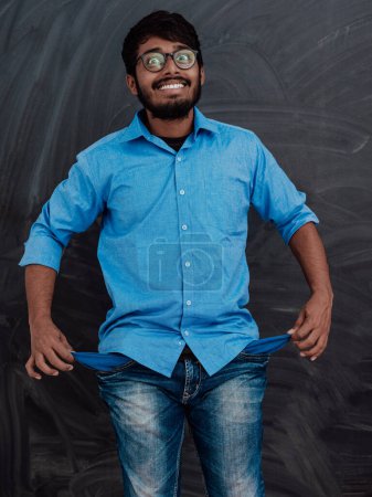 Photo for Photo of an Indian man wearing a blue shirt and glasses while symbolically showing his empty pockets on the background of a school blackboard - Royalty Free Image