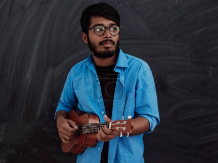Téléchargez les photos : Jeune homme indien en chemise bleue et lunettes jouant de la guitare devant le tableau noir de l'école. - en image libre de droit