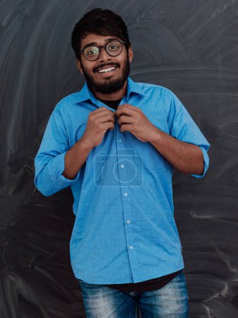 Foto de Joven estudiante indio con gafas se pone una camisa azul mientras está parado frente a la pizarra de la escuela. - Imagen libre de derechos