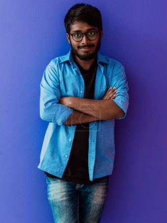 Foto de Un joven estudiante indio con una camisa azul con gafas posando con los brazos cruzados posando sobre púrpura. - Imagen libre de derechos