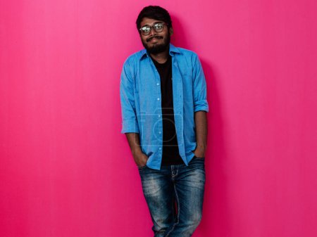 Foto de Hombre joven sonriente indio con camisa azul y gafas posando sobre fondo rosa. - Imagen libre de derechos