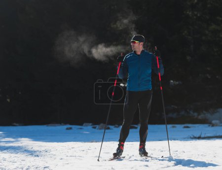 Foto de Retrato apuesto atleta masculino con esquís de fondo, tomando aliento fresco y teniendo un descanso después de un duro entrenamiento en un bosque cubierto de nieve. Concepto saludable de estilo de vida invernal. Foto de alta calidad - Imagen libre de derechos