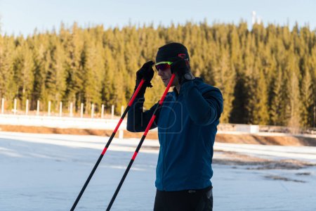 Téléchargez les photos : Beau athlète masculin avec des skis de fond préparant l'équipement pour l'entraînement dans une forêt enneigée. Je vérifie les montres intelligentes. Mode de vie sain en hiver. Photo de haute qualité - en image libre de droit