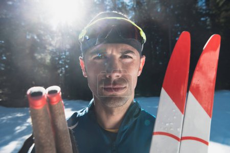 Téléchargez les photos : Portrait bel athlète masculin avec des skis de fond dans les mains et les lunettes, l'entraînement dans la forêt enneigée. Concept de mode de vie hivernal sain. Photo de haute qualité - en image libre de droit