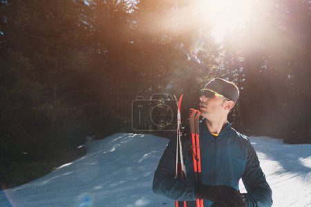 Téléchargez les photos : Portrait bel athlète masculin avec des skis de fond dans les mains et les lunettes, l'entraînement dans la forêt enneigée. Concept de mode de vie hivernal sain. Photo de haute qualité - en image libre de droit