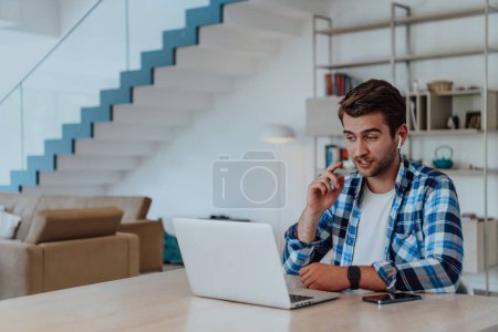 Photo for The man sitting at a table in a modern living room, with headphones using a laptop for business video chat, conversation with friends and entertainment. - Royalty Free Image