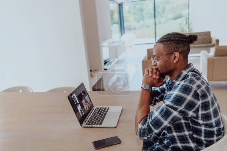 Photo for African American man in glasses sitting at a table in a modern living room, using a laptop for business video chat, conversation with friends and entertainment. - Royalty Free Image