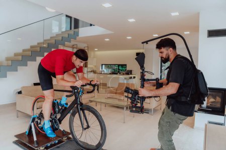 Foto de Un camarógrafo filmando a un atleta montando una bicicleta de triatlón en una máquina de simulación en una sala de estar moderna. Formación en condiciones pandémicas - Imagen libre de derechos
