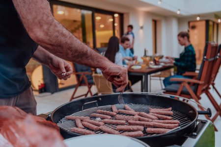Téléchargez les photos : Un groupe d'amis et barbecue en famille ensemble le soir sur la terrasse devant une grande maison moderne. - en image libre de droit