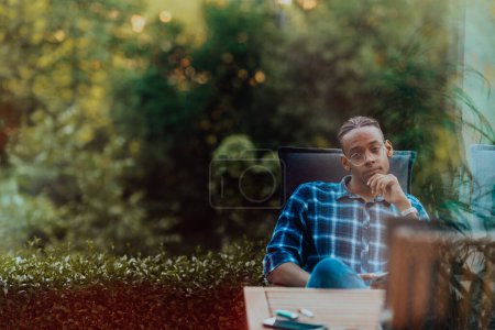 Téléchargez les photos : Un Afro-Américain assis devant la maison sur une terrasse moderne et regarde attentivement la caméra. - en image libre de droit