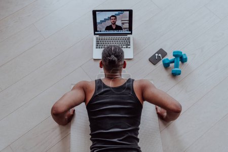 Foto de Entrenamiento en casa. Hombre deportivo haciendo el entrenamiento mientras ve un tutorial en línea en un ordenador portátil, haciendo ejercicio en la sala de estar, espacio libre. - Imagen libre de derechos